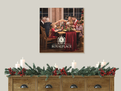 Wooden wall sign featuring a family Christmas dinner scene with the Royal Place logo, displayed above a festive candle and garland arrangement.