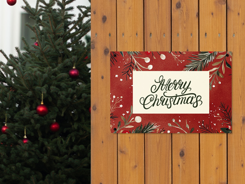 Red Merry Christmas dibond sign with greenery accents mounted on a wooden wall next to an outdoor Christmas tree.